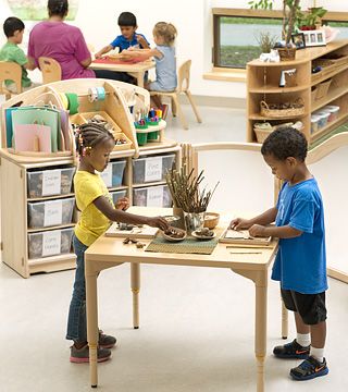 Two children are doing an art project while standing at a table in a Montessori-style classroom