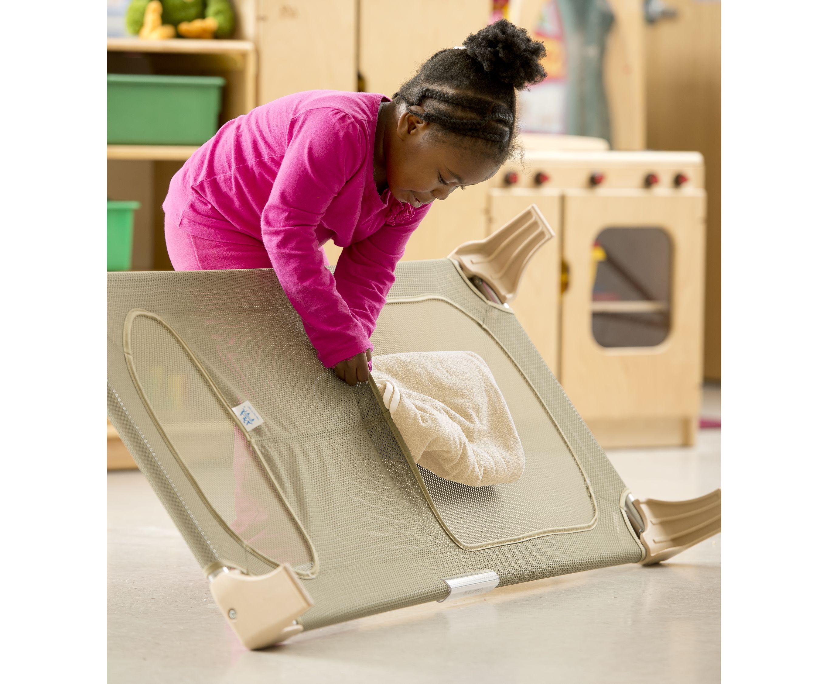 A young child in a pink shirt places a light-colored blanket into the mesh storage pocket of a beige cot.