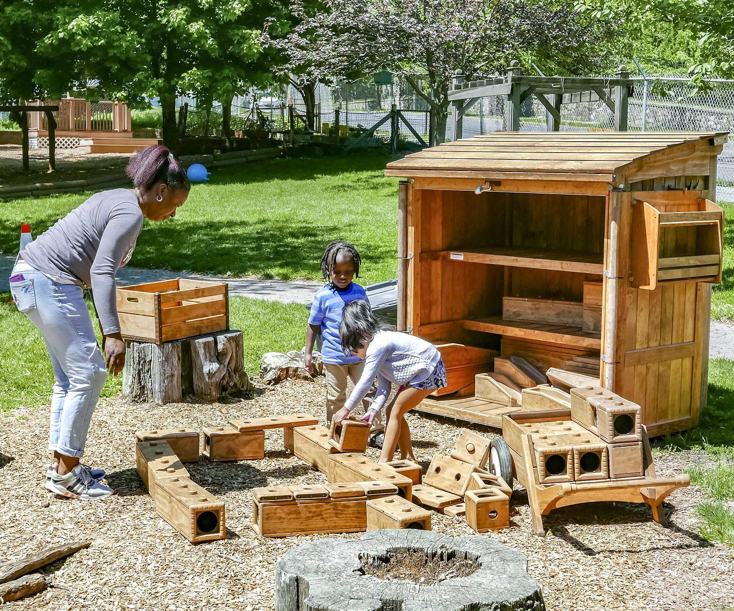 Adult and two children playing with large wooden Outlast blocks outdoors, next to a wooden storage shed.