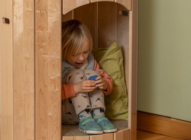 A young child with blonde hair sits curled up inside a wooden calming nest, holding a blue object.