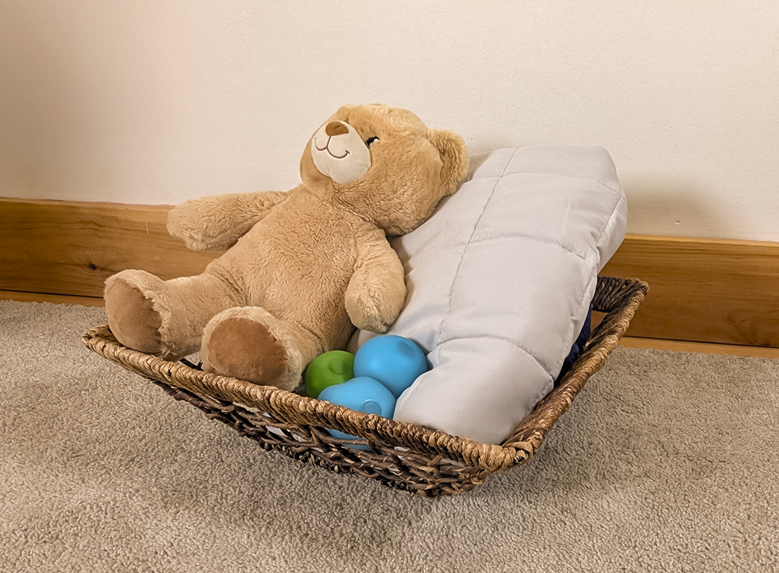 A basket with a teddy bear, pillow and stress ball in it.