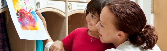 An adult and a child look at a colorful watercolor painting together, with cubbies in the background.